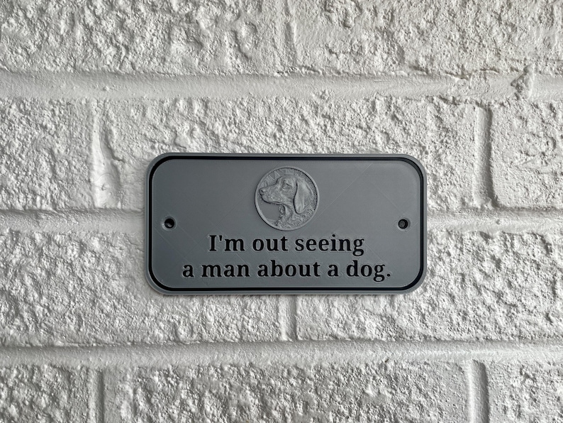 May include: A rectangular, gray metal sign with rounded corners is mounted on a white brick wall. The sign features a dog silhouette in a circle and the phrase, "I'm out seeing a man about a dog."