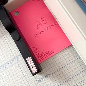 May include: A bright pink A5 cutting mat with the text A5 148mm x 210mm embossed in the center. The mat features decorative corner flourishes and is positioned within a paper trimmer with a black arm labeled TOOL BOX and a white grid base.