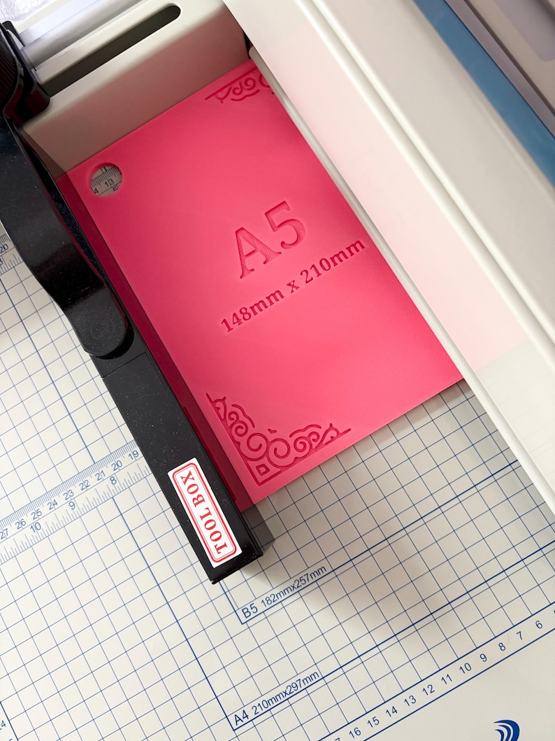 May include: A bright pink A5 cutting mat with the text A5 148mm x 210mm embossed in the center. The mat features decorative corner flourishes and is positioned within a paper trimmer with a black arm labeled TOOL BOX and a white grid base.