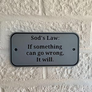 May include: A rectangular, silver-colored sign with rounded corners is mounted on a white brick wall. The sign has black text that reads "Sod's Law: If something can go wrong, It will."