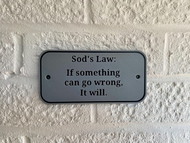 May include: A rectangular, silver-colored sign with rounded corners is mounted on a white brick wall. The sign has black text that reads "Sod's Law: If something can go wrong, It will."