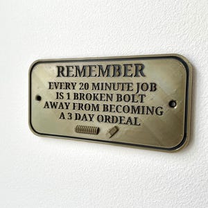 May include: A gold-colored metal sign with the text "REMEMBER EVERY 20 MINUTE JOB IS 1 BROKEN BOLT AWAY FROM BECOMING A 3 DAY ORDEAL".