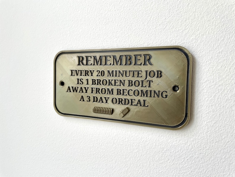 May include: A gold-colored metal sign with the text "REMEMBER EVERY 20 MINUTE JOB IS 1 BROKEN BOLT AWAY FROM BECOMING A 3 DAY ORDEAL".