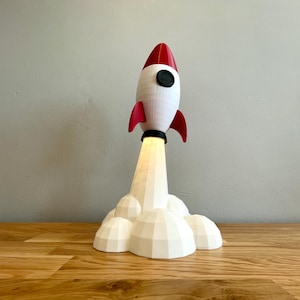May include: A white and red rocket ship lamp with a black porthole and red fins. The rocket is launching from a cloud-shaped base, with a warm light emanating from the bottom. The lamp is on a wooden surface.