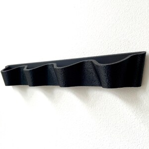 May include: A black, wall-mounted organizer with four wavy compartments. The organizer is made of a textured material and is mounted on a white wall. The design is simple and modern, suitable for various storage needs.
