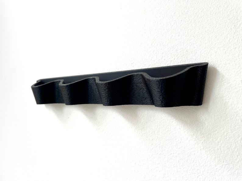 May include: A black, wall-mounted organizer with four wavy compartments. The organizer is made of a textured material and is mounted on a white wall. The design is simple and modern, suitable for various storage needs.