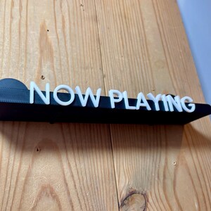 May include: A black sign with white letters that reads "NOW PLAYING".