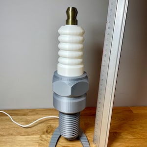 Oversized spark plug lamp with the white ceramic section designed as a light shade. Bronze top section, white shade and silver base. With a white cable. Stood on a wooden desk and grey background.
