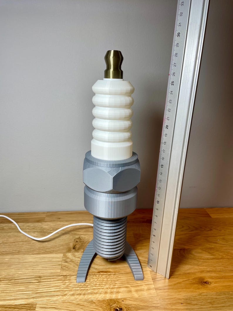 Oversized spark plug lamp with the white ceramic section designed as a light shade. Bronze top section, white shade and silver base. With a white cable. Stood on a wooden desk and grey background.