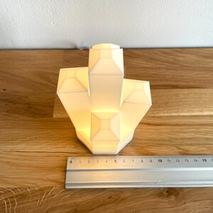 Pu&ograve; includere: Una lampada bianca a forma di cristallo geometrico con un bagliore caldo. La lampada &egrave; appoggiata su una superficie di legno.