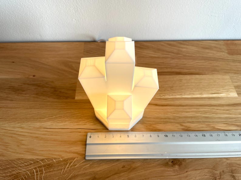 Pu&ograve; includere: Una lampada bianca a forma di cristallo geometrico con un bagliore caldo. La lampada &egrave; appoggiata su una superficie di legno.
