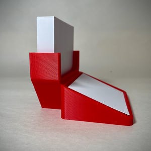 May include: A red 3D-printed desk organizer with a white angled surface and a vertical white block. The organizer is designed to hold items like notepads or small documents. The design is modern and minimalist.