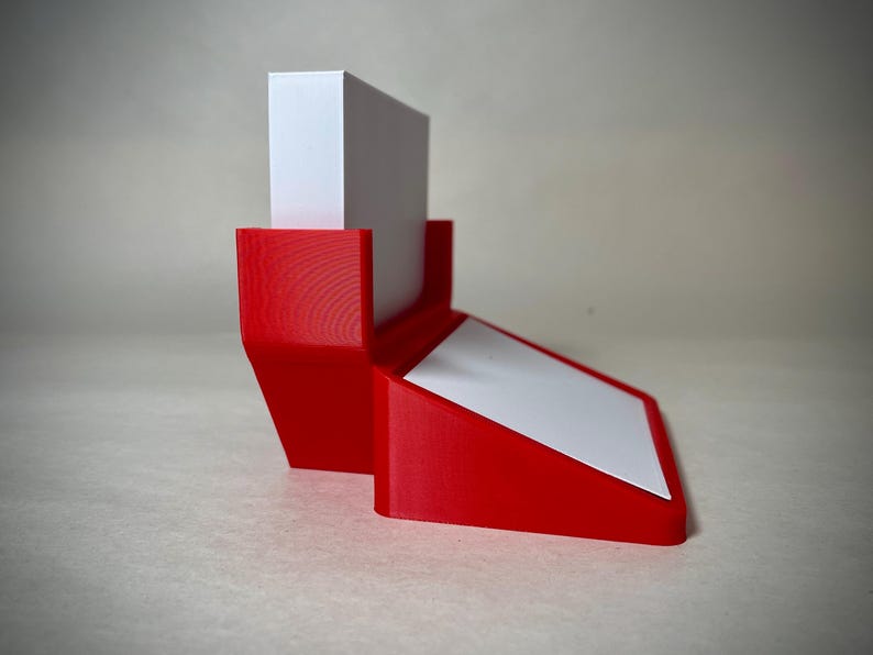 May include: A red 3D-printed desk organizer with a white angled surface and a vertical white block. The organizer is designed to hold items like notepads or small documents. The design is modern and minimalist.