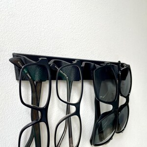 May include: A black eyeglasses holder mounted on a white wall, displaying several pairs of eyeglasses and sunglasses. The holder is designed to keep eyewear organized and accessible. The glasses have black frames.