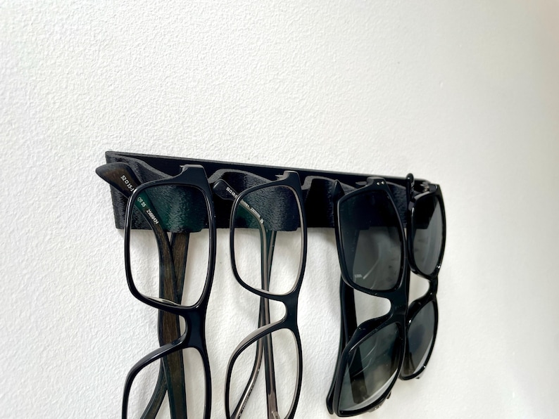 May include: A black eyeglasses holder mounted on a white wall, displaying several pairs of eyeglasses and sunglasses. The holder is designed to keep eyewear organized and accessible. The glasses have black frames.