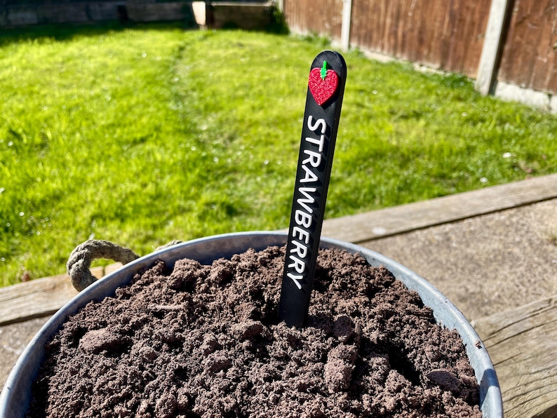 May include: A black plant marker with the word "STRAWBERRY" written on it. The marker has a red heart-shaped strawberry on top.