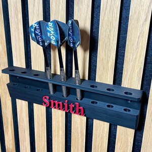 May include: A black dart holder with three darts, mounted on a wooden wall. The darts have blue and white flights with the word "POWER". The name "Smith" is in red on the holder.