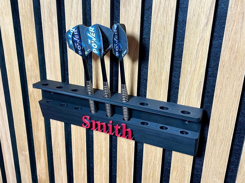 May include: A black dart holder with three darts, mounted on a wooden wall. The darts have blue and white flights with the word "POWER". The name "Smith" is in red on the holder.