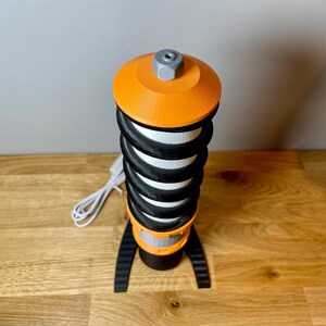 Op de afbeelding: Een zwart-oranje 3D-geprinte lamp met een spiraalvormig ontwerp en een wit snoer.