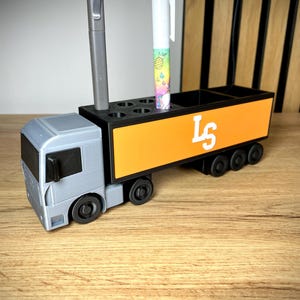 Pu&ograve; includere: Un organizer da scrivania grigio e arancione a forma di camion. Il camion ha una cabina grigia e un rimorchio arancione con le lettere "LS" in bianco. L'organizer contiene penne e matite.