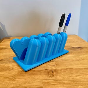 May include: A bright blue desk organizer featuring a series of cylindrical slots for pens and a heart-shaped end piece. The organizer is rectangular with a flat base and holds one black marker and one blue marker in the upright cylindrical openings.