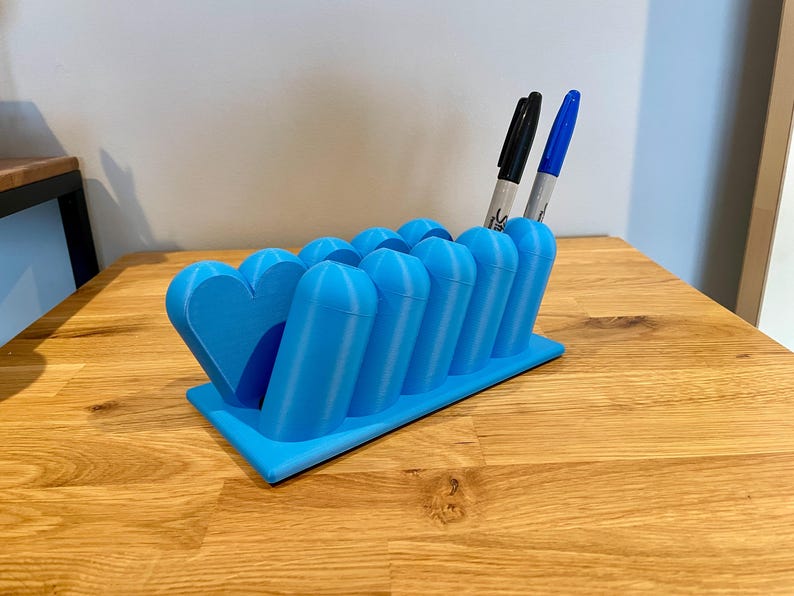 May include: A bright blue desk organizer featuring a series of cylindrical slots for pens and a heart-shaped end piece. The organizer is rectangular with a flat base and holds one black marker and one blue marker in the upright cylindrical openings.