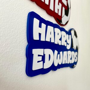 May include: Blue and red name plaques with white lettering. The blue plaque reads "Harry Edwards" with a soccer ball graphic. The red plaque is partially visible, with the name "Harry" in white. These are likely personalized items.