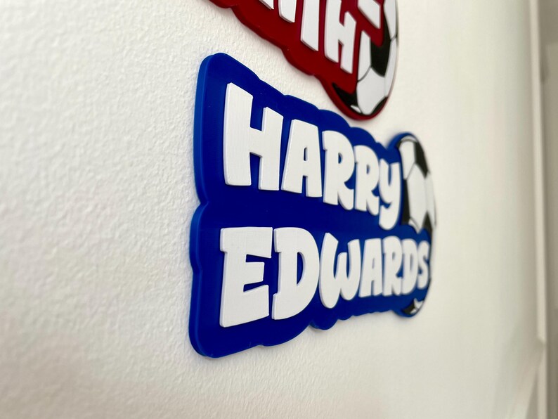 May include: Blue and red name plaques with white lettering. The blue plaque reads "Harry Edwards" with a soccer ball graphic. The red plaque is partially visible, with the name "Harry" in white. These are likely personalized items.