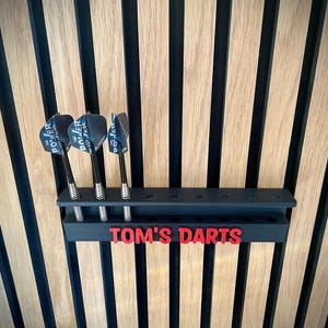 May include: A black dart holder with three darts, mounted on a wooden wall. The darts have black and silver shafts and blue and white flights. The holder has the text "TOM'S DARTS" in red.