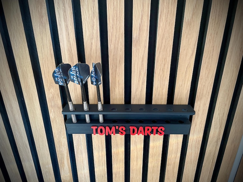 May include: A black dart holder with three darts, mounted on a wooden wall. The darts have black and silver shafts and blue and white flights. The holder has the text "TOM'S DARTS" in red.