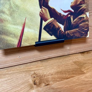 May include: A black metal poster hanger attached to a wooden wall, holding a poster featuring a person holding a spear, with a yellow and red background.