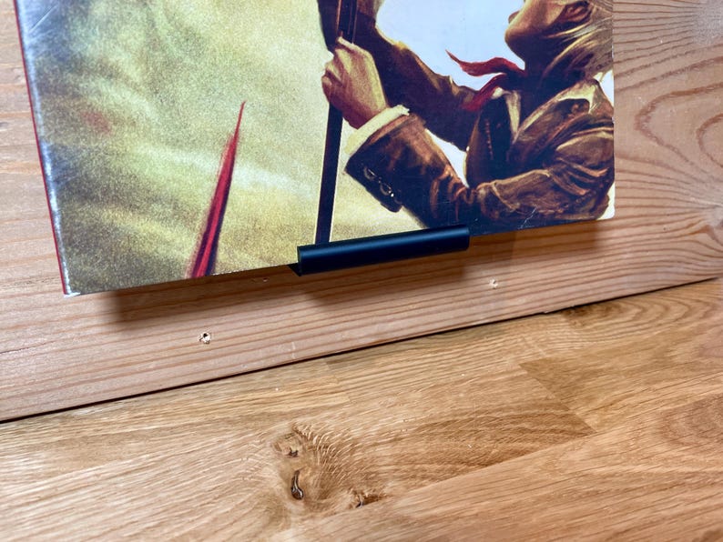 May include: A black metal poster hanger attached to a wooden wall, holding a poster featuring a person holding a spear, with a yellow and red background.