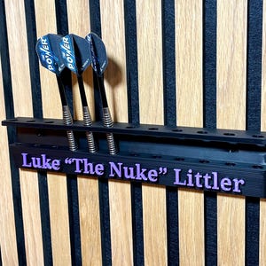 Puede incluir: Soporte negro para dardos con tres dardos. Los dardos tienen ca&ntilde;as negras y plumas azules y negras. El soporte tiene el nombre "Luke 'The Nuke' Littler" en letras moradas.
