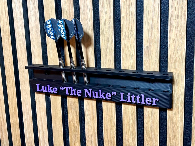 Puede incluir: Soporte negro para dardos con tres dardos. Los dardos tienen ca&ntilde;as negras y plumas azules y negras. El soporte tiene el nombre "Luke 'The Nuke' Littler" en letras moradas.