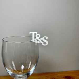 Wine glass with a set of initial letters clipped onto the side. Drink topper is white and the photo shows a grey background.