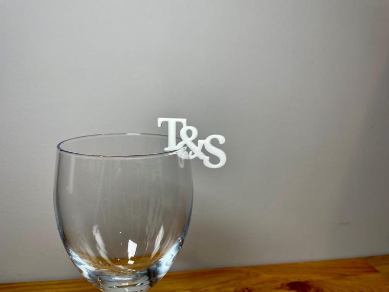 Wine glass with a set of initial letters clipped onto the side. Drink topper is white and the photo shows a grey background.