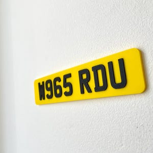 May include: A rectangular yellow sign with black lettering that reads "W965 RDU". The sign is mounted on a white textured wall. The sign is a license plate.