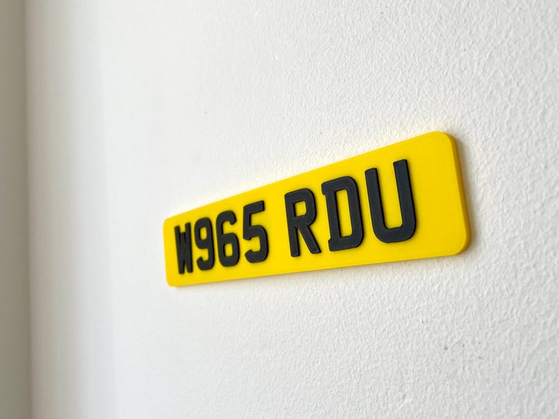 May include: A rectangular yellow sign with black lettering that reads "W965 RDU". The sign is mounted on a white textured wall. The sign is a license plate.