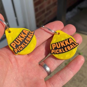 May include: Two yellow keychains with the text "PUKKA PICKLEBALL" in black and orange. The keychains are teardrop-shaped with a silver key ring attached. The background is a hand holding the keychains.