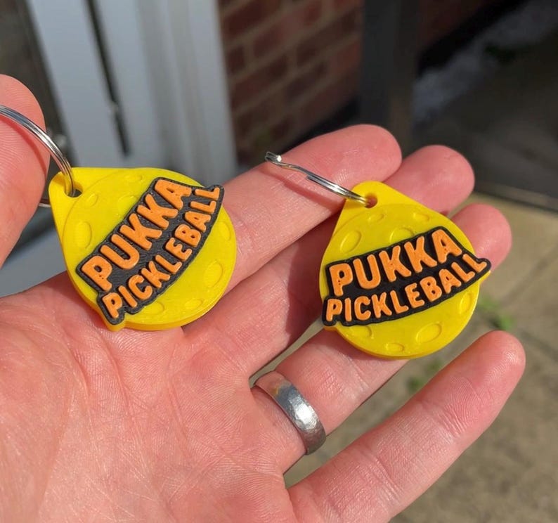 May include: Two yellow keychains with the text "PUKKA PICKLEBALL" in black and orange. The keychains are teardrop-shaped with a silver key ring attached. The background is a hand holding the keychains.