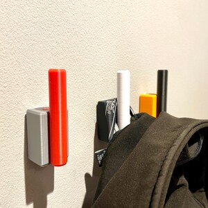Op de afbeelding: Drie wandhaken met een zwarte, witte en rode cilindrische pen. Een zwart jack hangt aan de witte pen.