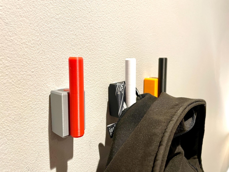 Op de afbeelding: Drie wandhaken met een zwarte, witte en rode cilindrische pen. Een zwart jack hangt aan de witte pen.