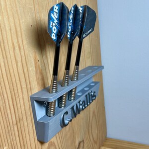 Silver / grey wall mounted dart holder with large black text along the lower edge showing a name. 3 darts in the holder with a total of 6 available slots. Shown mounted to a wooden background.