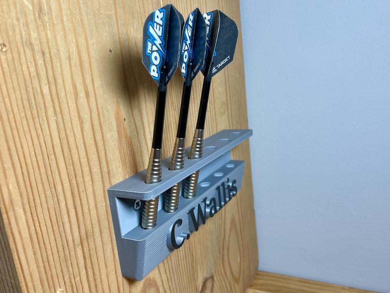 Silver / grey wall mounted dart holder with large black text along the lower edge showing a name. 3 darts in the holder with a total of 6 available slots. Shown mounted to a wooden background.