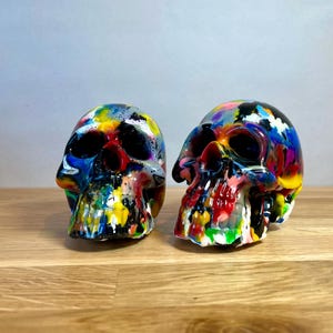 May include: Two colourful, hand-painted human skulls with black, white, red, yellow, blue, and green paint splatters. The skulls are made of resin and are approximately 15 cm tall.