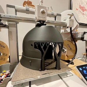 K&ouml;nnte beinhalten: Ein 3D-Drucker mit der Bezeichnung "Bambu Lab" druckt ein dunkelgraues, baumf&ouml;rmiges Objekt. Der Drucker hat einen wei&szlig;en Rahmen und ein strukturiertes Druckbett. Eine Filamentrolle und ein Tablet sind im Hintergrund zu sehen.