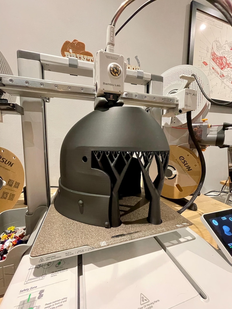 K&ouml;nnte beinhalten: Ein 3D-Drucker mit der Bezeichnung "Bambu Lab" druckt ein dunkelgraues, baumf&ouml;rmiges Objekt. Der Drucker hat einen wei&szlig;en Rahmen und ein strukturiertes Druckbett. Eine Filamentrolle und ein Tablet sind im Hintergrund zu sehen.