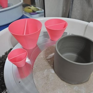May include: A pottery wheel setup featuring three pink, cone-shaped tools with dividers, and a grey ceramic cup. The tools are arranged on a silver surface, with a round, tan-coloured surface and a white basin in the background.