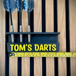 以下が含まれることがあります： 黄色い文字で「TOM'S DARTS」と書かれた、3本のダーツが入った濃い青色のダーツホルダー。ダーツは黒いシャフトと青と黒のフライトが付いています。黄色の巻尺が下に置かれています。