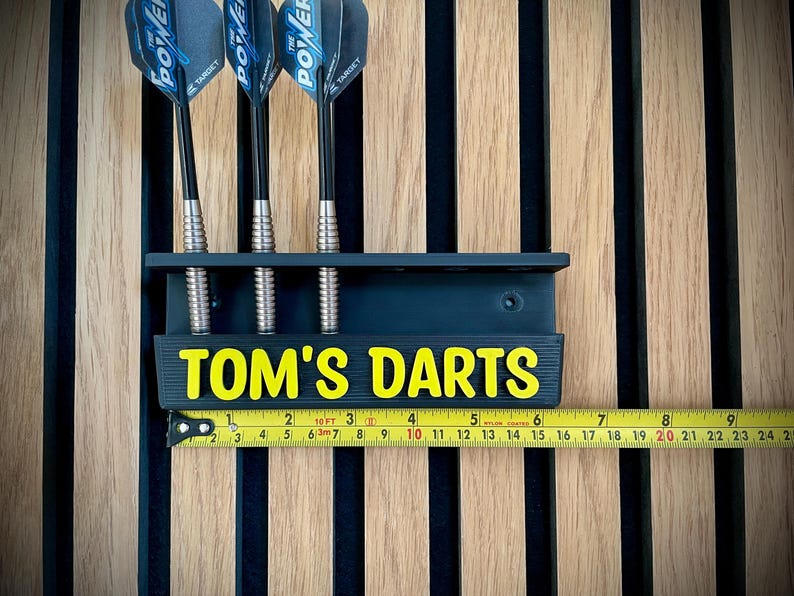 以下が含まれることがあります： 黄色い文字で「TOM'S DARTS」と書かれた、3本のダーツが入った濃い青色のダーツホルダー。ダーツは黒いシャフトと青と黒のフライトが付いています。黄色の巻尺が下に置かれています。